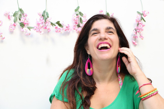 Luciana Peker. Fuente: Wikipedia Luciana Peker se retrata en una foto. Es una mujer de cabello cafe obscuro, y largo, más abajo de sus hombros. Está sonriendo con vista hacía al horizonte y arriba, mano levantada a la mejilla de su cara, con sonrisa grande y boca abierta. Viste una camiseta verde con aretes grandes y pulseras de muchos colores. En el fondo contra pared hay seis ramitas de flores color rosa pálida con hojas verdes, colgadas cada rama de manera deocrativa, hojas y pétalos hacía abajo.